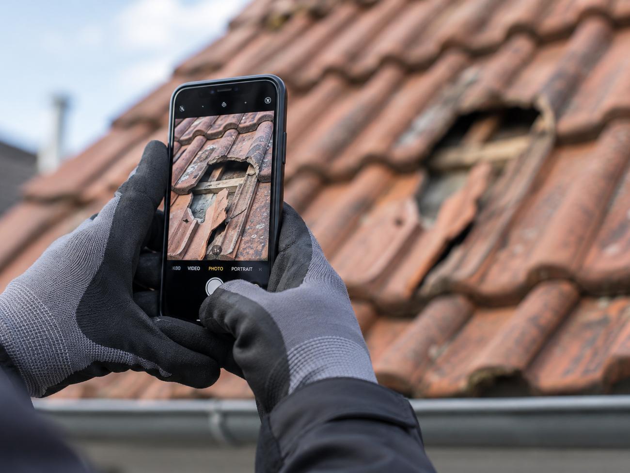 Behandschuhte Hände fotografieren mit dem Smartphone eine schadhafte Stelle im Ziegeldach